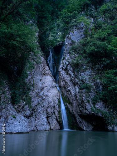 A tall waterfall falls from a cliff into a clear lake at night, lit by the light of the moon.