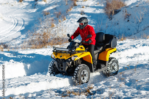 Attractive young man in red warm winter clothes and black helmet on the ATV 4wd quad bike stand in heavy snow with deep wheel track. Moto winter sports.