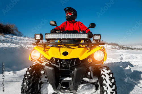 Photo of attractive young man in red warm winter clothes and black helmet on the ATV 4wd quad bike stand in heavy snow with deep wheel track. Moto winter sports.