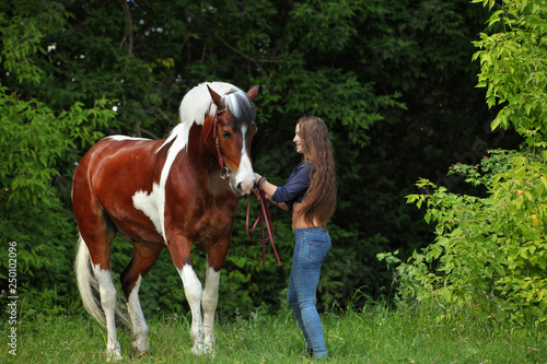 Beautiful model girl leading her horse in woods glade at sunset