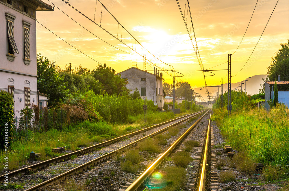 Railway tracks with old buildings on sides, wires above and Tuscany hills and mountains with dramatic cloudy yellow orange sky background, evening, dusk, twilight view, near Lucca town, Italy