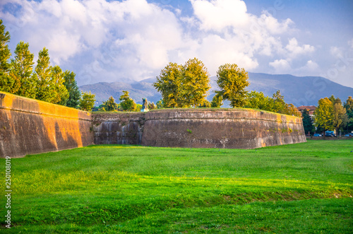 Defensive brick city wall, grass green lawn, trees and Tuscany hills and mountains with beautiful cloudy evening sky background, Lucca, Italy