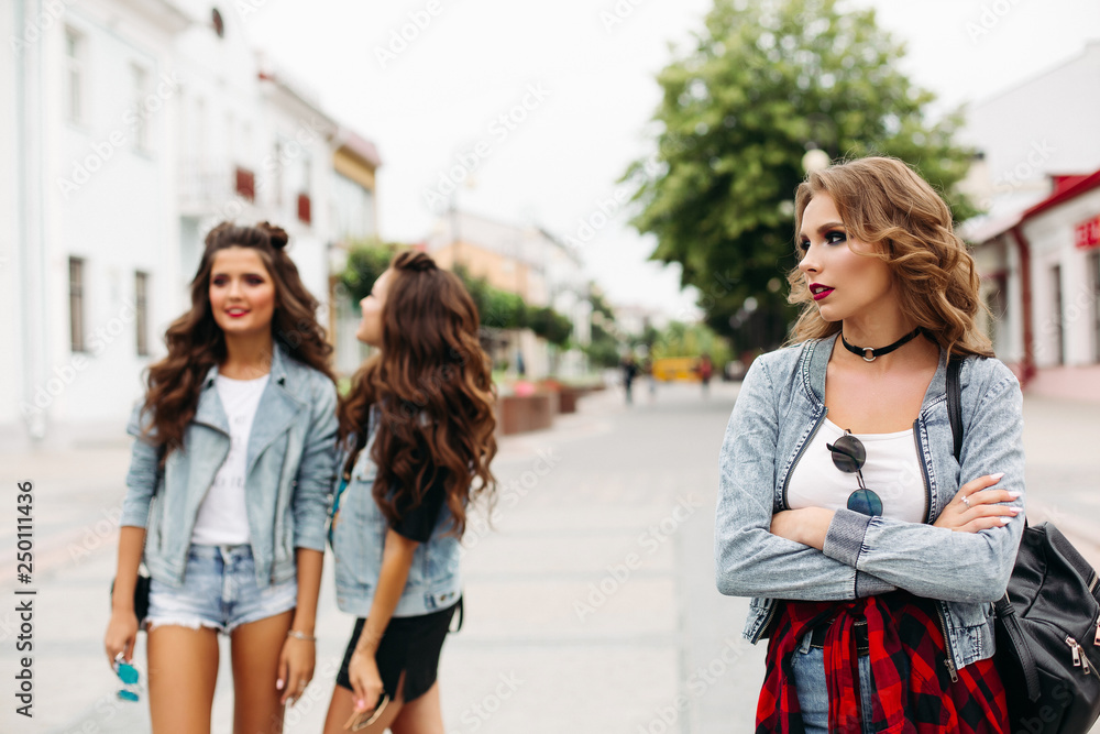 Portrait of beautiful sad girl with blonde wavy hair and bright make up with folded arms looking away against two fashionable best friends smiling and discussing her in the street. Blurred background.