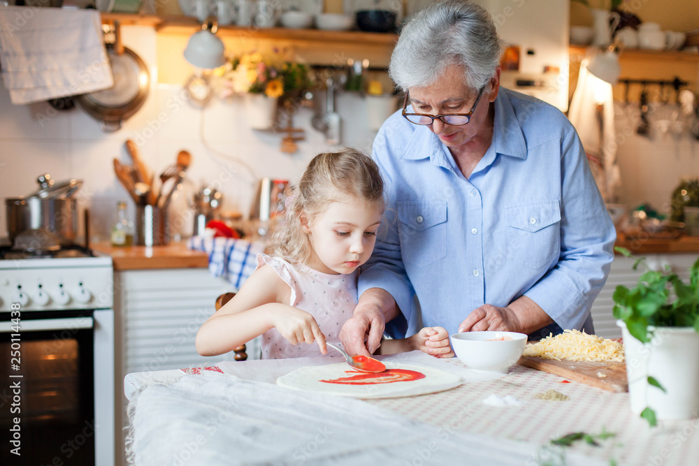Kid and grandmother are cooking italian pizza in cozy home kitchen for family dinner. Cute girl is helping to prepare homemade food. Old senior woman is teaching child. Children chef concept. Kid and grandmother are cooking italian pizza in cozy home kitchen for family dinner. Cute girl is helping to prepare homemade food. Old senior woman is teaching child. Children chef concept.