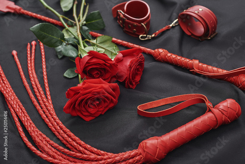 Playing BDSM games. Top view of bdsm leather kit (handcuffs, whip) and roses against of black silk fabric photo