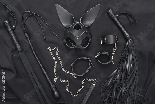 Explore the world of kinky play. Top view of bdsm leather kit (whips, handcuffs, mask and chain) against of a black silk.