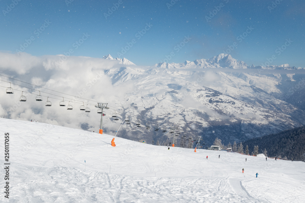 Unidentified skiers on a ski slope in the winter resort La Plagne in French Alps. The highest European mountain Mont Blanc above clouds in background.