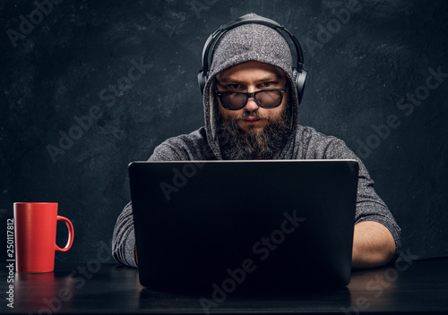 A hacker sits behind a laptop in a dark room and stares intently at the camera