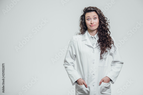 Horizontal portrait of a young pretty doctor's girl. Smiling, looking straight into the camera. On a gray background, holding hands in his pocket and pointing to a copy of the space