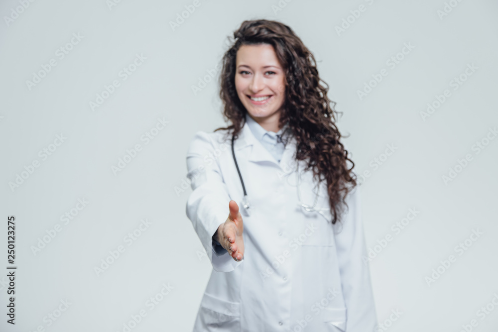 Portrait of happy young smiling girl doctor. Dressed in a white robe. Standing flat, one hand puts straight ahead in front of the gray background. Portrait of happy young smiling girl doctor. Dressed in a white robe. Standing flat, one hand puts straight ahead in front of the gray background.
