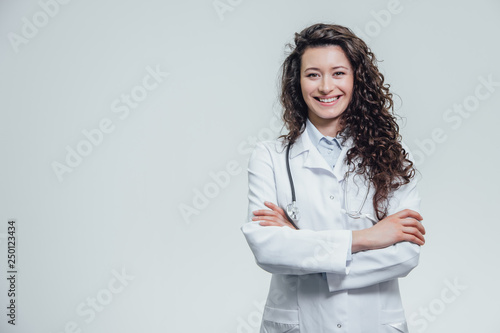 Portrait of happy young smiling girl doctor. Dressed in a white robe. Evenly standing with crossed hands on a gray background.