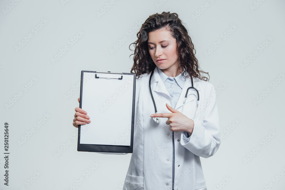 A young woman doctor is on a gray background. During this, it holds a folder with documents. Showing one of the items. A young woman doctor is on a gray background. During this, it holds a folder with documents. Showing one of the items.