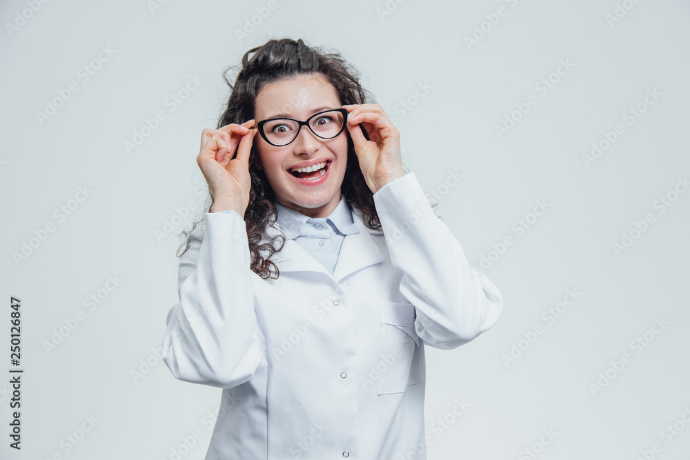 Beautiful smiling woman - a doctor in glasses, looking at the camera half the turn. Happy and funny woman wearing a medical front portrait over a gray background. Beautiful smiling woman - a doctor in glasses, looking at the camera half the turn. Happy and funny woman wearing a medical front portrait over a gray background.