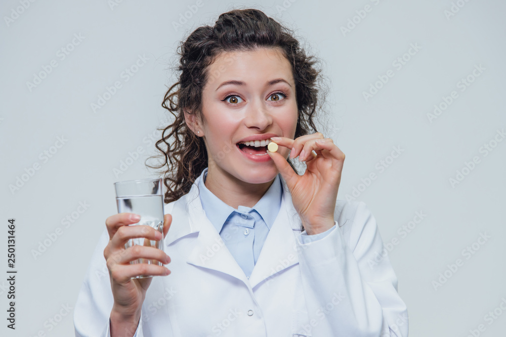 A young girl doctor takes vitamins for bones. At the same time, she stands on a gray background dressed in a white robe. Holds a glass of water and a white pill in his hands. A young girl doctor takes vitamins for bones. At the same time, she stands on a gray background dressed in a white robe. Holds a glass of water and a white pill in his hands.