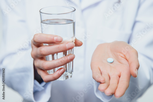 A young girl doctor takes vitamins. At the same time, she stands on a gray background dressed in a white robe. Holds a glass of water and a pill in his hands.