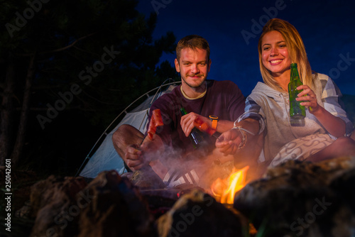 Young couple baking sausages on the campfire and drinking beer in the forest hill in the dusk