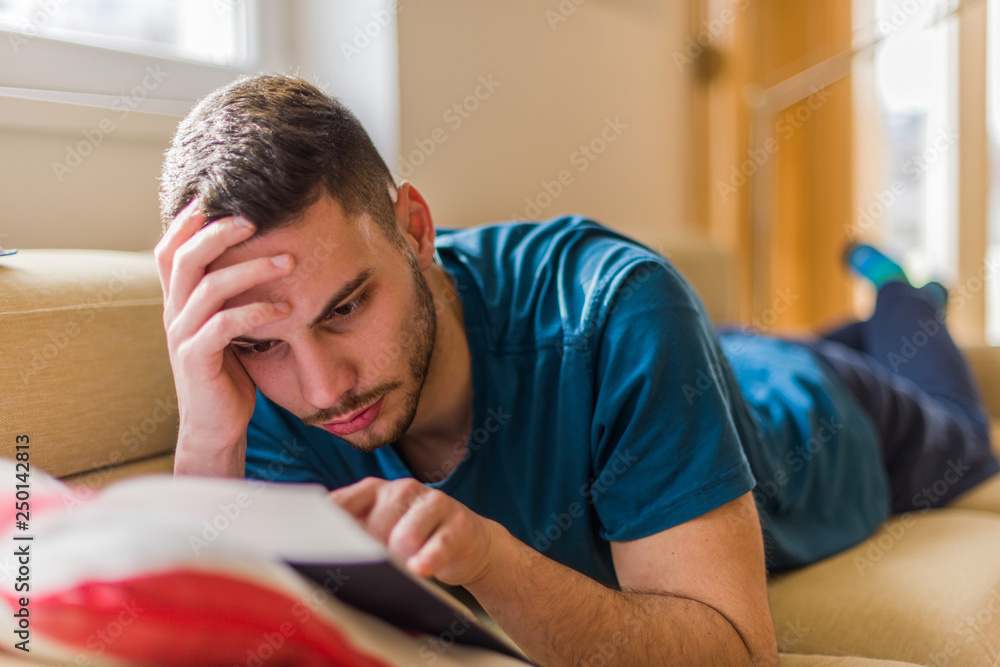 Worried man learning for exam from book on sofa Worried man learning for exam from book on sofa