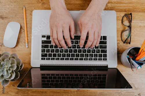 Top View of trendy wooden Office Desk with keyboard, white earphones and office supplies, working mans hands.