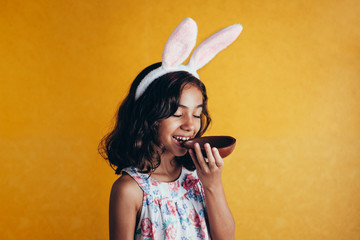 Cute little child wearing bunny ears on Easter day on color background. Girl eating chocolate easter egg