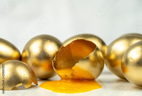 Golden eggs on white background, one egg broke, orange yolk flows out of Golden shell