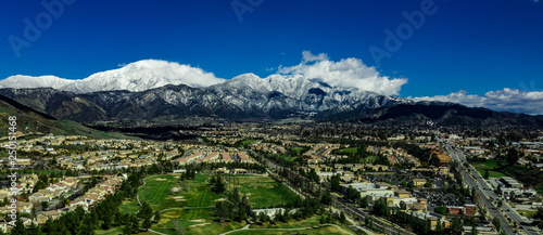 Panoramic, drone view of snow covered Mount San Gorgonio and the Little San Bernardino Mountains above Yucaipa Valley with white clouds, blue sky and green hills