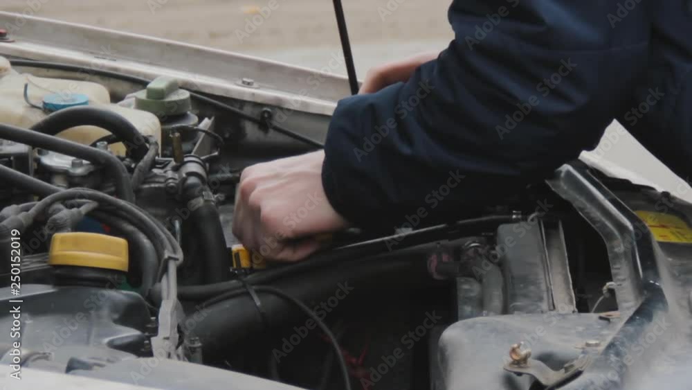 The driver fixes the malfunction of the car. Raised hood. View of the engine, battery and filler caps and black rubber pipes. Close-up of minor repairs. Men's hands with a tool in a winter jacket. The driver fixes the malfunction of the car. Raised hood. View of the engine, battery and filler caps and black rubber pipes. Close-up of minor repairs. Men's hands with a tool in a winter jacket.