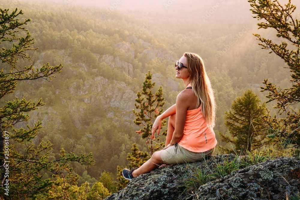 Naklejka premium beautiful young long-haired woman in a t-shirt and shorts sitting on a rock in the mountains in the summer.