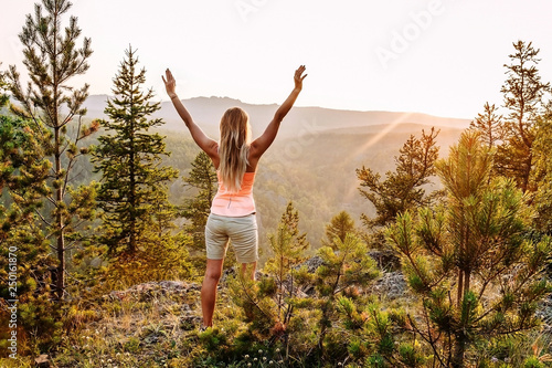 a slender young long-haired woman in a t-shirt and shorts stands with her hands up on the mountain