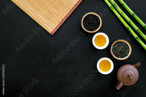 Chinese tea ceremony concept. Tea pot, tea cup, dry tea leaves, bamboo mat on black background top view copy space pattern