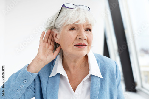 Come again please? Attractive mature woman wearing blue jacket and spectacles on her head eavesdropping. Stylish retired female in elegant clothes holding hand at her ear, suffering from hearing loss photo