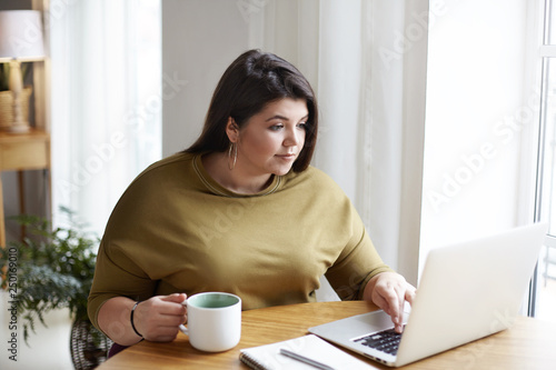 Chubby attractive young female freelancer wearing elegant sweater and round earrings working in front of open laptop, sitting in cozy home office interior, drinking coffee, browsing websites