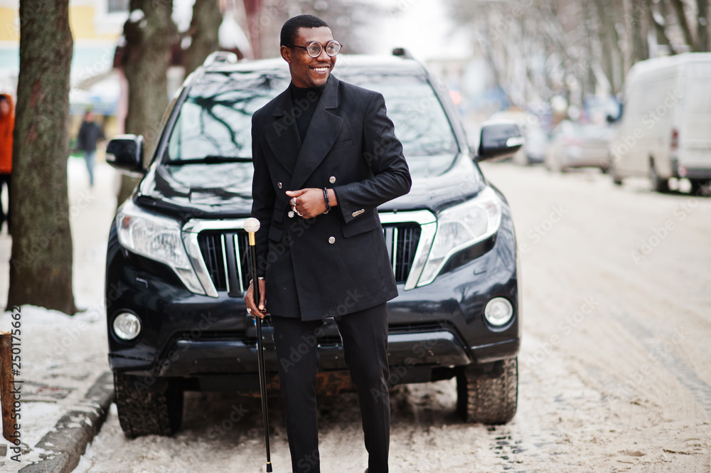 Stylish african american gentleman in elegant black jacket, holding retro walking stick with golden diamond ball handle. Rich fashionable afro man against business suv car.