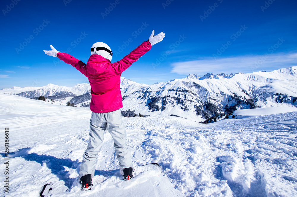 Snowy mountain view. Young happy woman snowboarder standing on the top of the mountain rising arms to the blue sky
