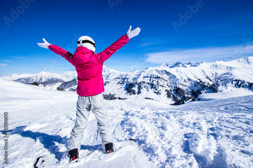 Snowy mountain view. Young happy woman snowboarder standing on the top of the mountain rising arms to the blue sky