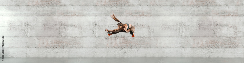 Sporty brunette dressed in sportswear and with ponytail jumping high. In background gray wall, copy space.