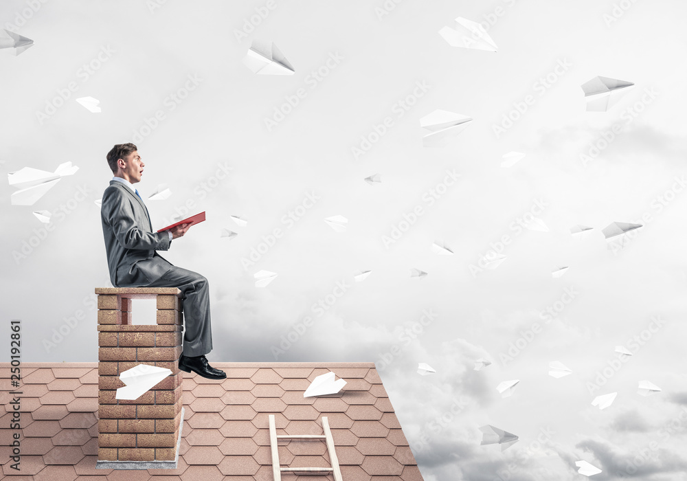 Man on brick roof reading book and paper planes flying in air Man on brick roof reading book and paper planes flying in air