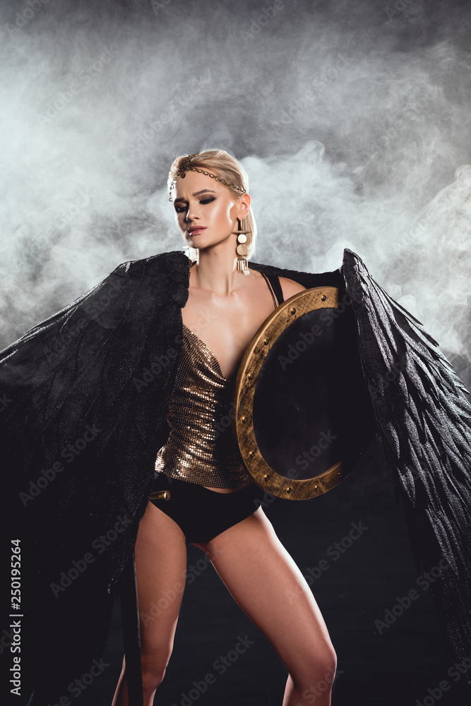 beautiful woman in warrior costume with black wings and shield posing on smoky background