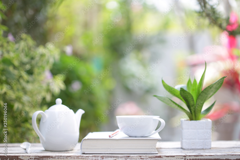 White coffee cup and kettle with notebook on wooden table with green plants