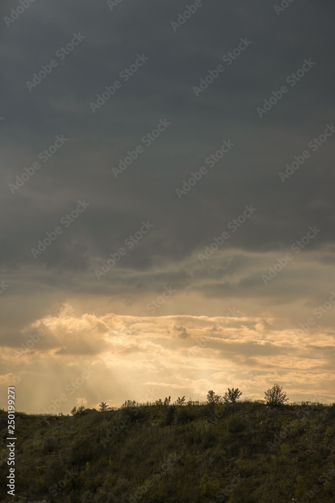 Spring landscape of grassy hill against a dramatic sunset sky with fluffy clouds and volumetric light rays passing through them Spring landscape of grassy hill against a dramatic sunset sky with fluffy clouds and volumetric light rays passing through them