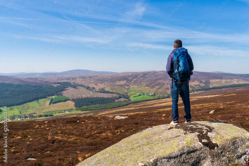 Tourist carrying a blue backpack standing on a large rock and admiring the beautiful view from the Two Rock Mountain peak in Dublin Mountains, Ireland. Irish hills landscape.