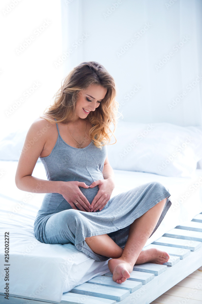 Beautiful pregnant young woman sits on a bed in a room and makes a heart on her tummy. Bright daylight sunlight from the windows Beautiful pregnant young woman sits on a bed in a room and makes a heart on her tummy. Bright daylight sunlight from the windows