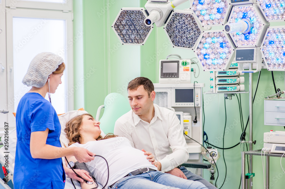 Obraz premium Beautiful pregnant woman with her husband in medical clinic. Nurse measuring pressure of pregnant woman