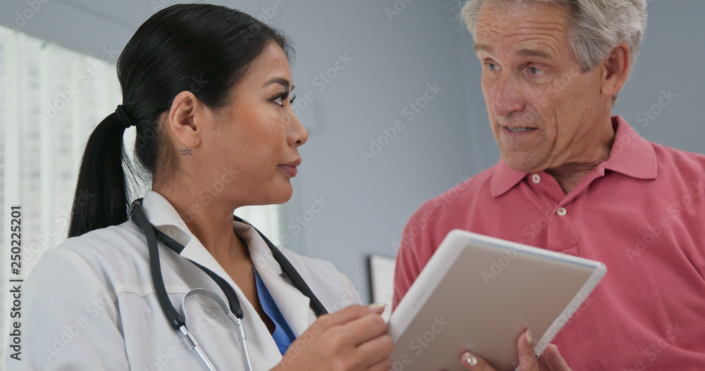 Low-angle view of Japanese woman primary care doctor using tablet computer while talking to senior Caucasian male patient. Physician taking history from aging man Low-angle view of Japanese woman primary care doctor using tablet computer while talking to senior Caucasian male patient. Physician taking history from aging man