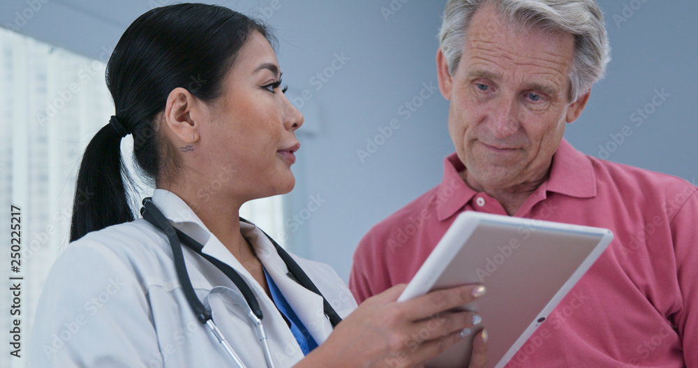 Japanese woman primary care doctor using tablet computer while explaining treatment to senior Caucasian male patient. Low-angle view of physician talking to aging man Japanese woman primary care doctor using tablet computer while explaining treatment to senior Caucasian male patient. Low-angle view of physician talking to aging man