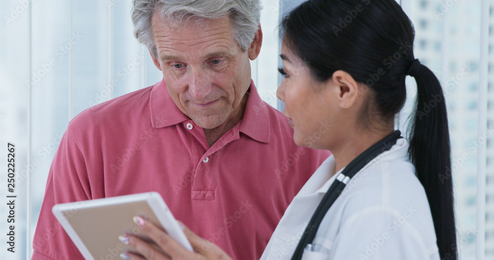 Elderly Caucasian male patient asking Japanese woman doctor questions while looking at tablet computer together. Female primary care physician and her patient discussing treatment Elderly Caucasian male patient asking Japanese woman doctor questions while looking at tablet computer together. Female primary care physician and her patient discussing treatment