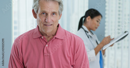 Portrait of senior Caucasian male patient looking at camera and smiling while doctor works in background. Attractive older man in hospital with female physician out of focus behind him