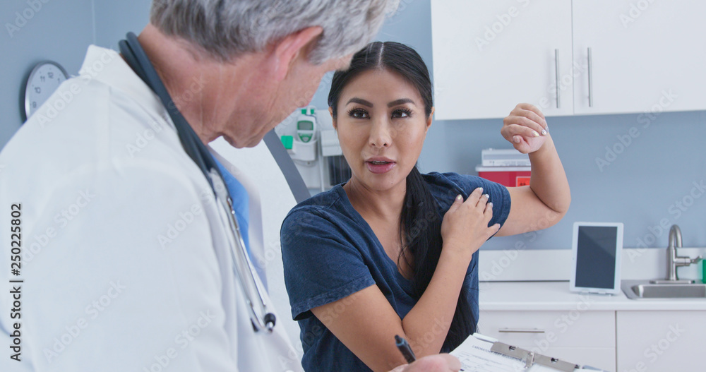 Japanese woman explaining shoulder pain to doctor in exam room. Patient with rotator cuff injury talking to medical professional Japanese woman explaining shoulder pain to doctor in exam room. Patient with rotator cuff injury talking to medical professional