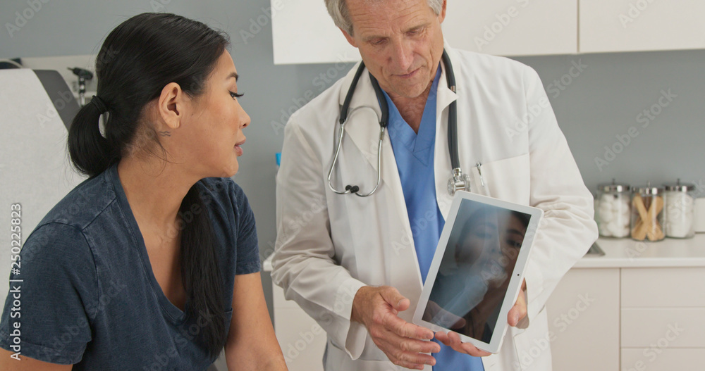 Asian woman sitting on exam table looking at tablet computer with knee x-ray as doctor explains treatment of injury. Senior medical professional using device with patient Asian woman sitting on exam table looking at tablet computer with knee x-ray as doctor explains treatment of injury. Senior medical professional using device with patient