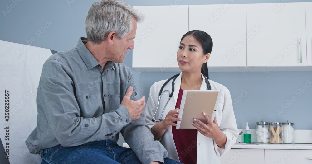 Obraz premium Asian woman doctor talking senior Caucasian male patient sitting on exam table. Medial professional using tablet computer to take medical history from older man
