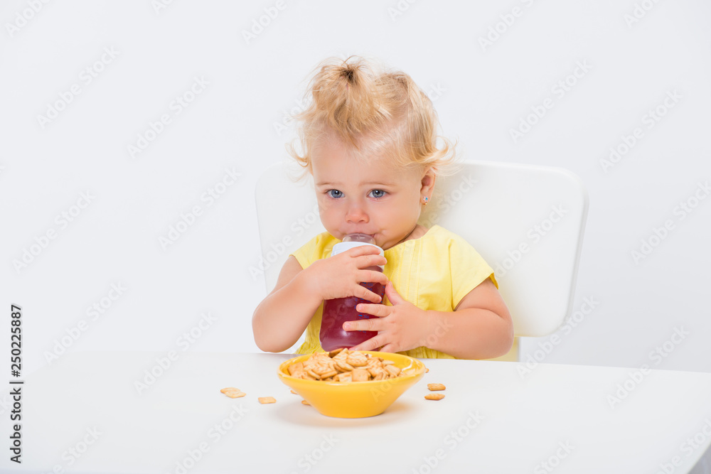 Cute little baby girl 1 year old eating cereal flakes and drinking juice or compote from a bottle at the table isolated on white background Cute little baby girl 1 year old eating cereal flakes and drinking juice or compote from a bottle at the table isolated on white background